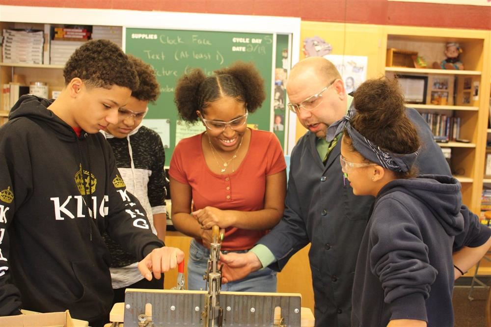 Students stand with teacher at a wood shop table. Students stand with teacher at a wood shop table.